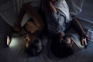 Young couple using their smartphones while lying back to back on bed at night