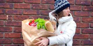Young woman goes with paper bag of vegetables and food from grocery store on brick wall background. Girl with paper package of food