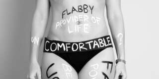 A woman's torso is shown in a black bikini bottom. Words are written on her skin that say 'flabby', 'provider of life', 'fat', 'powerful', 'ugly', 'beautiful' and 'uncomfortable?'. The image illustrates a woman's striggle with body image. It is in black and white.