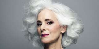 Mature woman with medium length, big, wavy, silvery, grey hair in front of grey background wearing make up red lip stick, portrait.