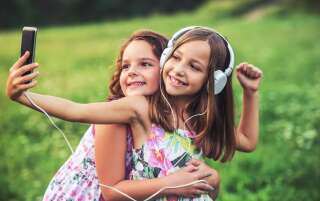Childhood time. Cute smiling little girls enjoying in the park while making a selfie and listening to music with mobile phone