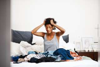 Shot of an unhappy young woman getting dressed with piles of clothing on her bed