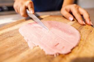 Cette photographie présente une personne en train de découper des morceaux de jambon sur une planche en bois à l'aide d'un couteau.