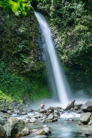 Tourist sitting on a rock in a river in front of majestic waterfall, Arenal, La Fortuna, Costa Rica