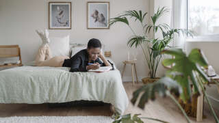 Black woman writing in a notebook while laying on a comfortable bed at home.