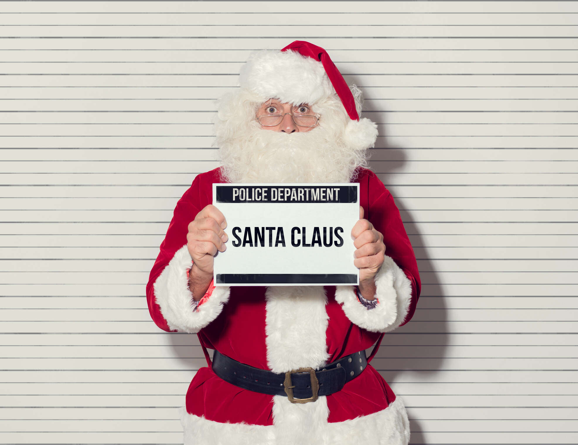 Santa Claus arrested on Christmas eve, he is posing for his mug shot at the police department and holding an identification board