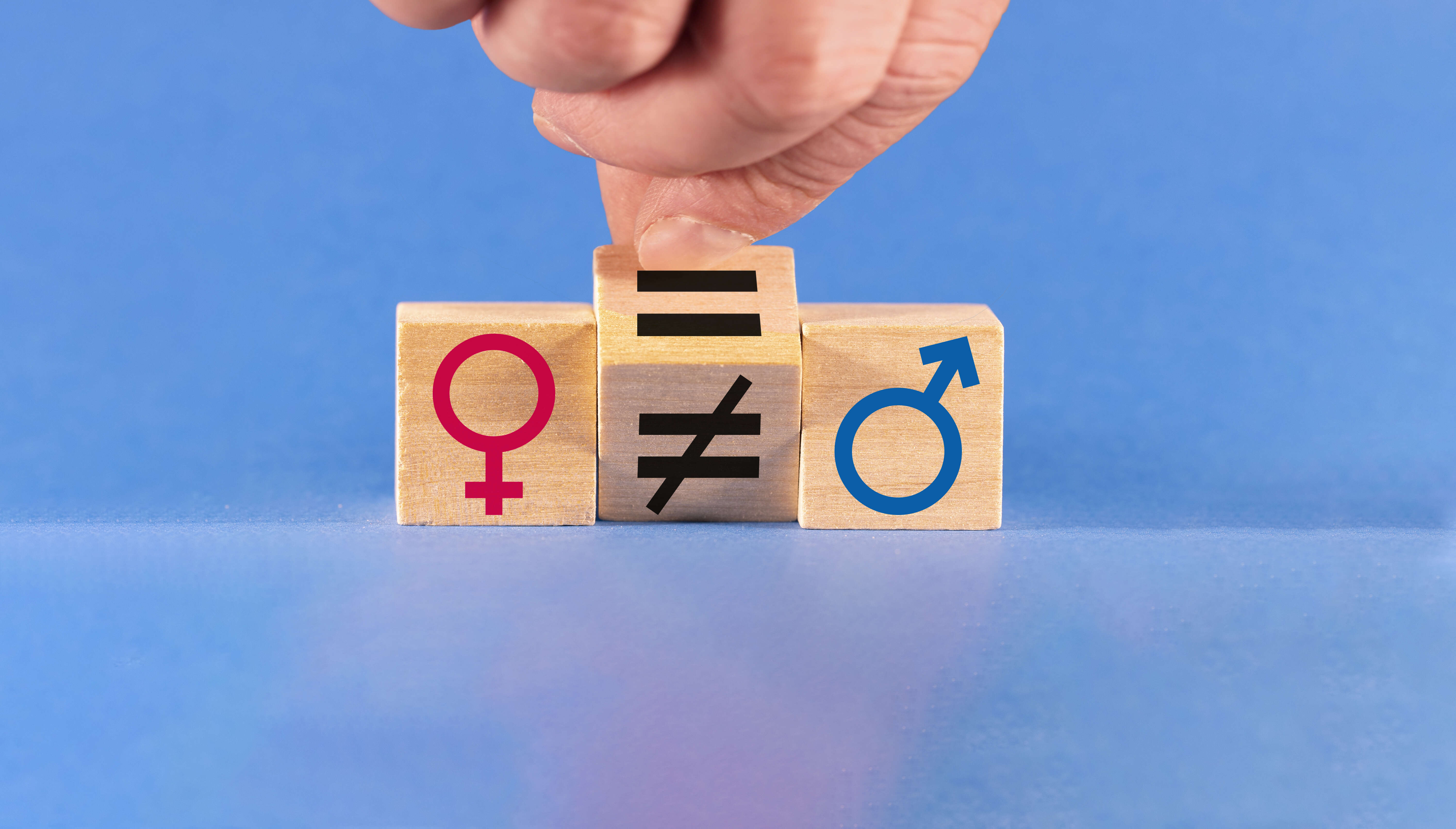 A hand turns a wooden die to change an unequal sign to an equal sign between male and female symbols on a blue background.