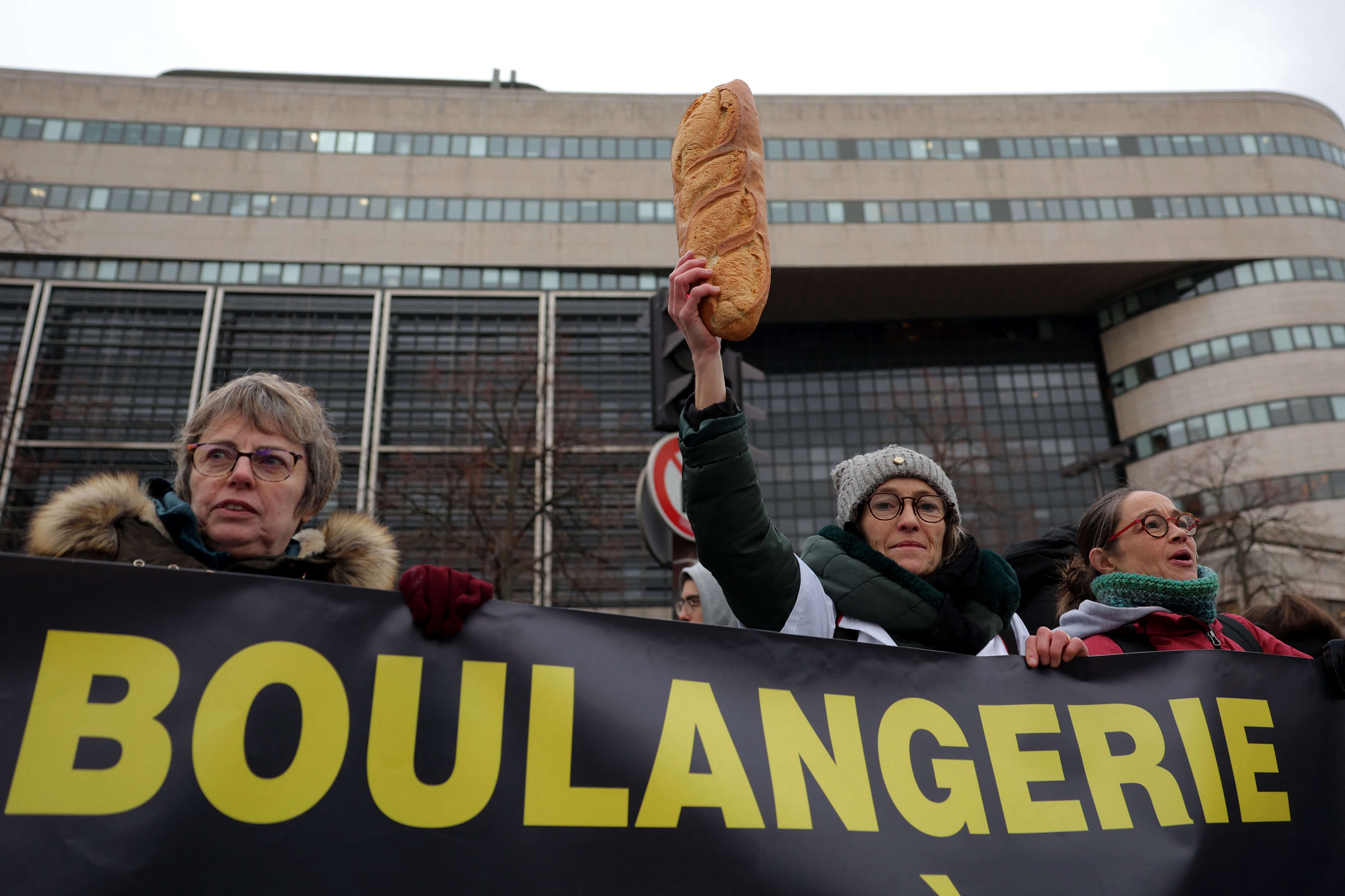 A woman holds a loaf of bread up as she stands behimd a sign that reads, 'bakery', during a protest organised by the collective for the survival of bakers and crafts artisans, at Bercy, in Paris on January 23, 2023. - Recently described as 