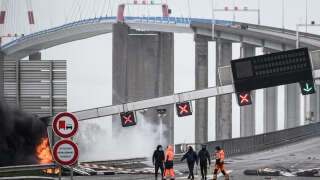 Protestors walk next to a damaged and burning road sign gantry at the entrance of the 
