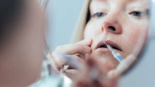 Woman in the mirror doing botox injection in lips.