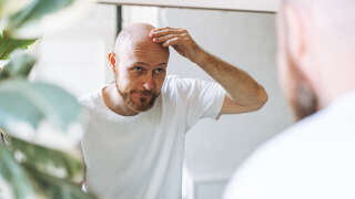 Young adult bearded man looking in mirror in bathroom touching head worried about about hair loss