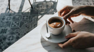 Close up of woman's hand holding a cup of coffee, drinking coffee in outdoor cafe against beautiful sunlight, having a relaxing moment. Enjoying life's simple pleasures