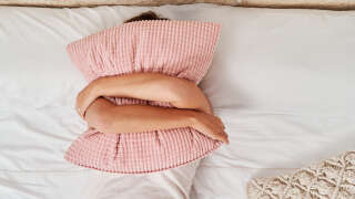 Top view of a beautiful young woman lying in bed covering her face with a pillow.