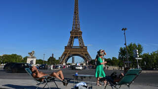 Passers-by walk past two people enjoying the sun in a long chair in front of the Eiffel Tower in Paris on August 21, 2023, as the red heatwave vigilance is activated in four departments, a sign of a 