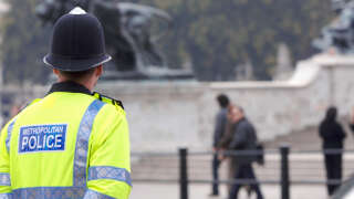 A British police man stands on duty across the road from Buckingham Palace, as tourists pass by in the background on their way to see the Changing of the Guard.
