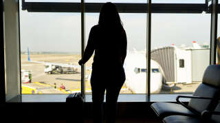 Against the light of a long-haired woman, with her luggage in front of the window, watching the plane ready to board at the airport