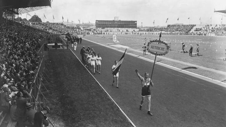 An Austrian athlete carrying the 'Autriche' placard leads the flagbearer and the Austrian team, gives the Olympic salute during the opening ceremony of the 1924 Summer Olympics, held at the Stade Olympique de Colombes, in the Colombes suburb of Paris, France, 5th July 1924. A variant of the Roman salute, the Olympic salute fell out of use due to its similarity to the Nazi salute. (Photo by Central Press/Hulton Archive/Getty Images)