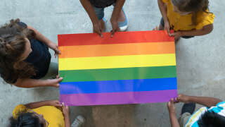 A group of multi-ethnic elementary students stand around a colorful gay pride poster.  They are each holding an edge of the poster so it is out flat for the camera to see.  Only the tops of their heads can be seen as the picture is taken from an aerial view.  Each of the students are wearing casual clothing.